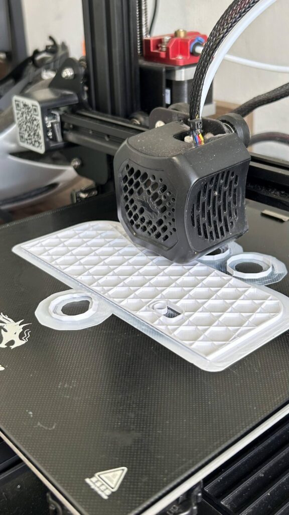 impressora 3D com filamento branco imprimindo uma base retangular. tem círculos em volta, um na frente e dá pra ver dois atrás, onde crescerão os suportes para as partes que cairiam sem eles. deve ter 1cm de altura por enquanto.
